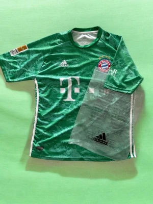 2019/2020 Bayern Munich Neuer Shirt  - Snygg grön målvaktströja från FC Bayern München med Neuer och nummer 1 på ryggen. Tröjan har vita detaljer, Adidas-logga och klubbmärke på bröstet samt sponsortryck. Tillverkad i ett lätt och ventilerande material, perfekt för fotbollsträning eller match.