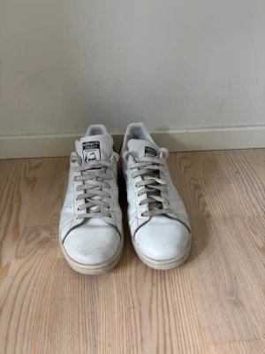 Vita Adidas Stan Smith sneakers - Klassiska vita Adidas Stan Smith sneakers med lågt skaft och svarta detaljer på hälen. Skorna har perforerade ränder på sidorna och är tillverkade i skinn med snörning framtill. Perfekta för en clean och tidlös stil.