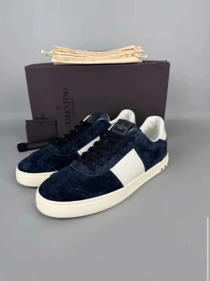 Valentino blå mocka sneakers - Snygga sneakers från Valentino i mörkblå mocka med vita detaljer och svart snörning. Yttersulan är vit med coolt kamouflagemönster undertill. Skorna har en klassisk låg profil och stilren design som passar till många outfits.