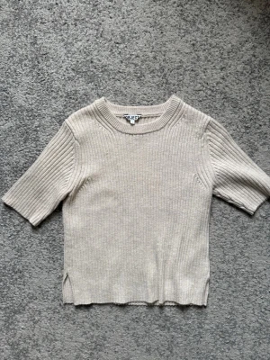 Beige ribbstickad topp från OBJECT - Säljer en beige ribbstickad topp från OBJECT med korta ärmar och rund hals. Tröjan har en rak passform, diskreta slitsar i sidorna och är gjord i ett mjukt, stretchigt material. Perfekt för en clean och enkel look.