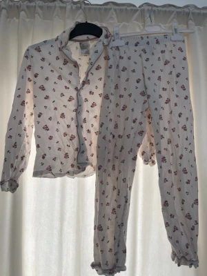 Blommig pyjamas med volangdetaljer barn  - Söt tvådelad pyjamas i ljusbeige med små rosa blommor. Skjortan har knappar framtill, krage och volangkant vid ärmslut och nederkant. Byxorna har resår i midjan och volang vid benslut. Perfekt för mysiga kvällar hemma. Finns några fläckar skriv för mer bilder 