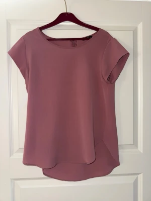 Rosa topp från ONLY - Säljer en enkel och stilren rosa topp från ONLY i storlek 36. Toppen har rund halsringning, korta ärmar och något längre bakstycke för en snygg siluett. Perfekt att matcha med jeans eller kjol för en trendig look.