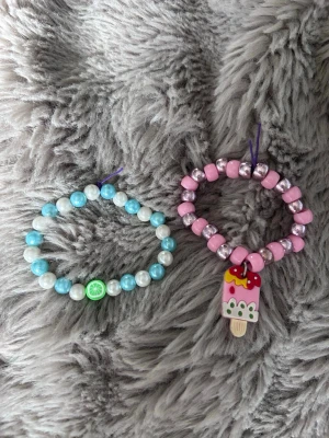 Två färgglada pärlarmband - Säljer två söta armband med pärlor. Ett armband har vita och turkosa pärlor med en limegrön citronskiva som detalj. Det andra har rosa och silverfärgade pärlor samt en glassberlock i rosa, vitt och gult med jordgubbsdekor. Perfekta för att piffa upp din stil!