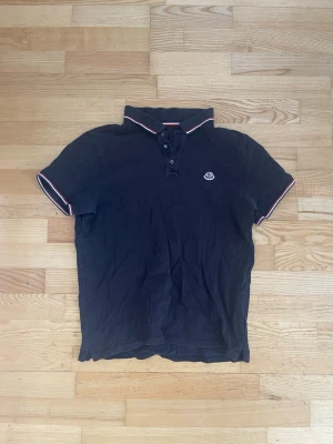 Moncler piké tröja - Svart äkta moncler piké tröja. Tröjan är i storlek large men sitter som small. Den är gammal och köpt begagnad för ca 2600 kr. Den har inga defekter förutomen att den har blivit lite urtvättad och tappat mörkheten i det svarta. 
