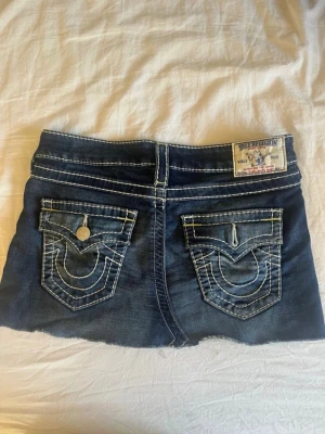 True Religion jeansshorts mörkblå denim - Säljer en kort jeanskjol från True Religion i mörkblå denim med kontrastsömmar och rå kant nertill. Kjolen har klassiska fickor fram och bak med dekorativa sömmar och knappdetaljer. Perfekt för dig som gillar streetwear och denim. Tyvärr är ena knappen av men den finns att sätta på.