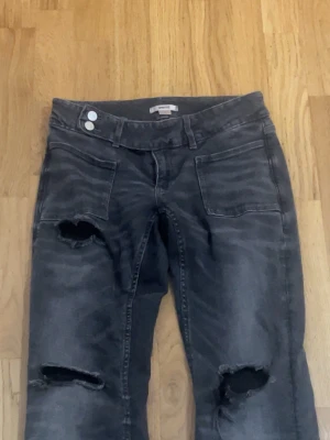 Grå lågmidjade jeans från Gina tricot - Säljer ett par grå jeans med slitningar och stora hål på båda benen. Jeansen har raka ben, två framfickor och dubbla knappar i midjan. Perfekta för en avslappnad och trendig streetstyle-look.