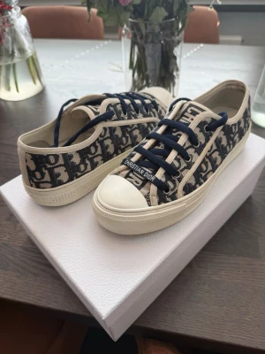 Dior Oblique sneakers med blå logga - Snygga Dior sneakers i storlek 38,5. De är väldigt sparsamt använda och är i gott skick, inga defekter förutom att texten i sulan har fadeat bort lite samt att ena skosnöret gått sönder lite vid toppen som syns i bild 5. De är köpta i butik så kvitto, dustbag och box tillkommer vid köp!