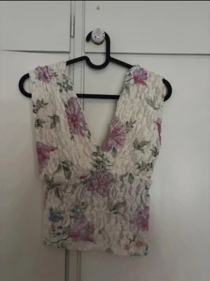 Blommig spets topp med v-ringning - Söt ärmlös topp i vit spets med blommigt mönster i rosa, lila och grönt. Djup v-ringning både fram och bak ger en romantisk vibe. Perfekt för varma dagar och somriga outfits.