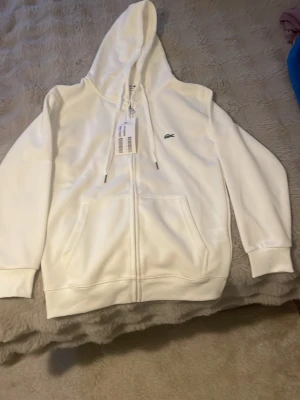 Lacoste zip hoodie  - Snygg vit hoodie från Lacoste med dragkedja framtill och klassisk krokodil-logga på bröstet. Hoodien har huva med dragsko och stora fickor framtill. Tillverkad i mjukt material som känns skönt mot huden. Perfekt för en clean och stilren look.