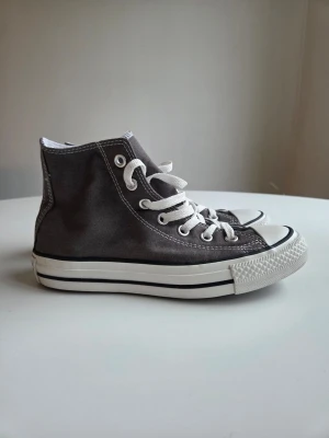 Grå Converse Chuck Taylor High - Klassiska Converse Chuck Taylor All Star high-top sneakers i grå canvas med vita gummisulor och vita skosnören. Ikonisk rund logga på sidan och metallöljetter för ventilation. Perfekt för en avslappnad och tidlös stil.