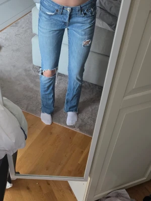 Ljusblå slitna jeans från True Religion - Säljer ett par ljusblå jeans från True Religion med raka ben och slitningar på låret och knät. Jeansen har klassiska detaljer på bakfickorna och låg midja. Perfekta för en avslappnad och trendig look.