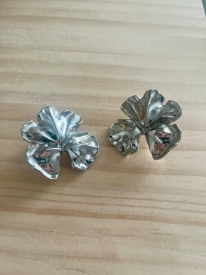 Stora silvriga blomörhängen från Lindex - Säljer ett par statementörhängen i form av stora blommor i blankt silverfärgat metallmaterial. Örhängena har en vågig, organisk form som ger en cool och modern look. Perfekta för dig som vill sticka ut med accessoarerna.