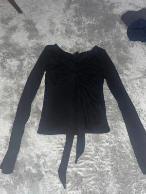 Knyt topp från Gina - Snygg svart långärmad topp med knyt framtill och lätt rynkade detaljer vid bysten. Mjuk och stretchig kvalitet som sitter skönt på kroppen. Perfekt till jeans eller kjol för en trendig look.