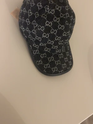 Mörkblå keps från Gucci - Snygg svart keps från Gucci i denim med det klassiska GG-mönstret över hela kepsen. Kepsen har böjd skärm och justerbar rem bak. Insidan är ljus och mjuk för extra komfort. Perfekt för dig som vill ha en trendig accessoar med lyxig känsla.