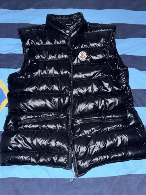 Svart dunväst från Moncler - Snygg svart dunväst från Moncler med glansig finish och hög krage. Västen har dragkedja framtill, två sidofickor med dragkedja och Moncler-logga på bröstet. Perfekt för lager-på-lager och streetwear-stil.
