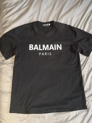 Svart Balmain Paris t-shirt - Snygg svart t-shirt från Balmain Paris med klassisk vit logga framtill. T-shirten har rund halsringning och korta ärmar. Tillverkad i mjuk bomull för en bekväm känsla och stilren look. PASSAR ÄVEN M.