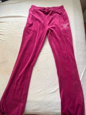 Rosa velourbyxor Juicy Couture - Mjukisbyxor i rosa velour från Juicy Couture med logga i vitt på höften. Byxorna har rak passform och resår i midjan för extra komfort. Perfekta för en chill och avslappnad stil.