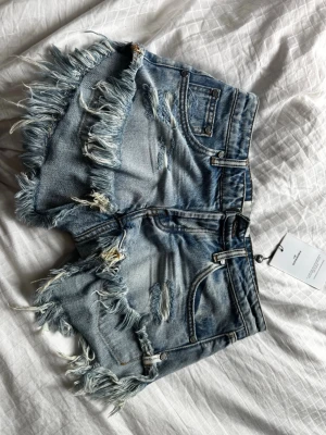 Blå fransiga jeansshorts från One Teaspoon - Säljer ett par blå denimshorts från one teaspoon med råa, fransiga kanter. Shortsen har slitna detaljer, fem fickor och klassisk knappgylf. Perfekta för sommaren och ger en avslappnad vibe. Jag har aldrig använt shortsen då de är försmå. 💕
