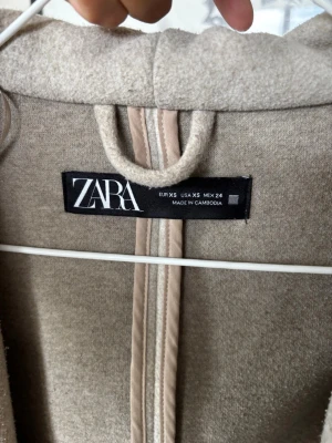 Beige kappa från Zara med stor krage - Stilren beige kappa från Zara i storlek XS. Kappan har en oversized krage och raka ärmar, samt en mjuk ull-liknande yta. Perfekt för dig som gillar minimalistisk och trendig design. Enkel att matcha med olika outfits.