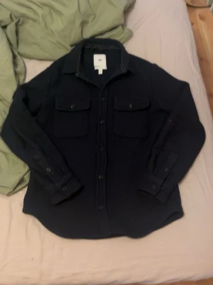 Mörkblå overshirt med fickor - Snygg mörkblå overshirt med två bröstfickor och knäppning framtill. Overshirten har klassisk krage och långa ärmar med knappar vid ärmsluten. Perfekt lager-på-lager-plagg för en avslappnad stil.