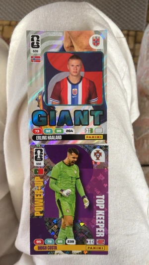 Panini FIFA samlarkort fotboll - Två Panini FIFA samlarkort med fotbollsspelarna Erling Haaland och Diogo Costa. Kortet med Haaland har holografisk effekt och texten 'GIANT', medan Costa-kortet har lila bakgrund och texten 'TOP KEEPER'. Båda korten har spelstatistik och landslagsflaggor.