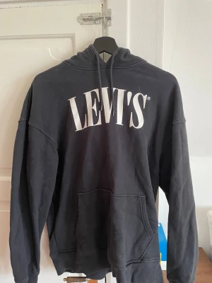 Svart hoodie från Levi's med logga - Säljer en svart hoodie från Levi's med stor vit logga på bröstet. Hoodien har huva med snörning, känguruficka framtill och långa ärmar. Perfekt för en chill och avslappnad stil.