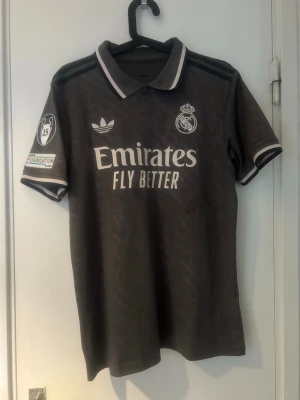 Real Madrid matchtröja Mbappé svart - Snygg svart Real Madrid matchtröja från Adidas med Mbappé och nummer 9 på ryggen. Tröjan har vita detaljer, klubbmärke och sponsortryck på bröstet samt Champions League-märke på ärmen. Perfekt för dig som älskar fotboll och vill sticka ut på planen eller läktaren.