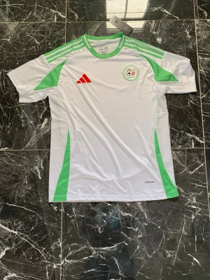 Algeriet fotbollströja Adidas vit/grön - Snygg vit fotbollströja från Adidas med gröna detaljer och Algeriets landslagsemblem på bröstet. Tröjan har röda Adidas-loggan, meshpaneler på sidorna för ventilation och klassiska Adidas-ränder på axlarna. Perfekt för träning eller match.