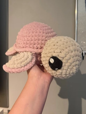 Handvirkad plushie  - Supergulligt handvirkat gosedjur i form av en sköldpadda. Sköldpaddan har ett mjukt beige huvud och ben, samt ett rosa skal. Stora svarta ögon med vita detaljer ger ett sött uttryck. Perfekt som ett gosedjur eller som en fin gåva!