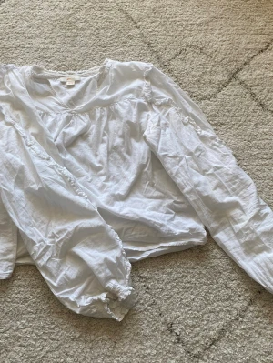 Vit blus med volang från Gina Tricot - Söt vit blus från Gina Tricot med långa ärmar och volangdetaljer längs axlar och ärmar. Blusen har en lös passform och resår vid ärmsluten. Tillverkad i ett mjukt och lätt tyg som känns skönt mot huden.