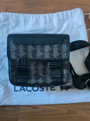 Svart axelväska med mönster från Lacoste - Snygg svart axelväska från Lacoste med geometriskt mönster i vitt och detaljer i läderimitation. Väskan har justerbar axelrem, två spännen framtill och orange innerfoder. Perfekt för att bära det viktigaste med stil.
