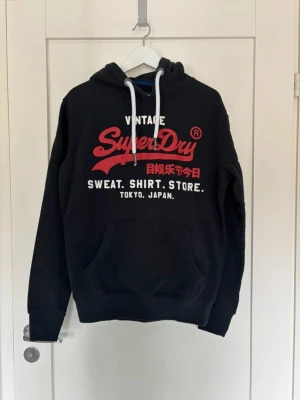 Superdry hoodie - Svart hoodie från Superdry med stor röd och vit logga och text på bröstet. Klassisk känguruficka framtill och vita dragsnören i huvan. Skön och avslappnad passform, perfekt till jeans eller joggers. Riktigt schysst hoodie i skit bra skick. Mitt pris 399 och priser är ej hugget i stenen så de e ba skicka bud!