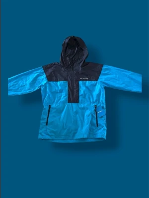 Blå och svart vindjacka Columbia - Snygg blå och svart vindjacka från Columbia med huva och halv dragkedja framtill. Jackan har två dragkedjeförsedda fickor och är tillverkad i lätt, vindtätt material. Perfekt för blåsiga dagar och outdoor-äventyr.