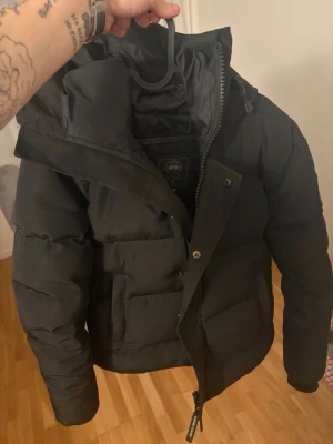 Svart pufferjacka från Canada Goose - Säljer en svart pufferjacka från Canada Goose i modellen Fusion S/P. Jackan har en hög krage, dragkedja med knappslå och stora fickor framtill. Perfekt för kalla vinterdagar och riktigt snygg till streetwear-stilen.