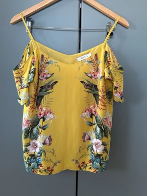 Gul blommig linnetopp från Oasis - Snygg gul linnetopp från Oasis med tunna axelband och blommigt mönster i grönt, rosa och vitt. Toppen har v-ringning och offshoulder-detalj med korta ärmar. Storlek 10.