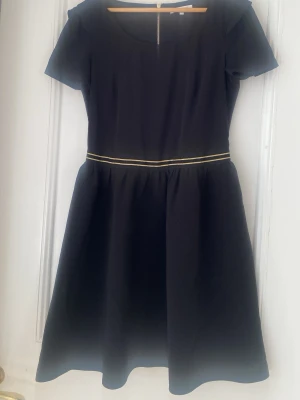 Svart klänning med gulddetalj - En svart klänning med rund halsringning och korta ärmar. Midjan markeras av ett tunt guldfärgat band som ger en snygg kontrast. Kjolen har en lätt utställd form och klänningen slutar strax under knät. Perfekt för dig som gillar enkel men elegant stil.