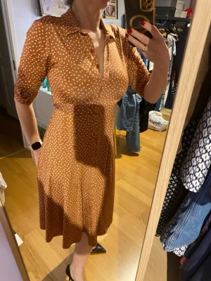 Brun prickig midiklänning med krage - Snygg ljusbrun klänning från Stockholms Klänningsfabrik med vita prickar, klassisk krage och knytband i midjan. Klänningen har trekvartsärm och v-ringad hals. Perfekt för dig som gillar retrovibba.