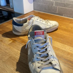 Golden Goose sneakers - Säljer ett par vita sneakers från Golden Goose med grå stjärndetalj och blå häl. Till skillnad från många GG-skor på Plick är dessa ÄKTA‼️ Skorna är i skick 7/10 skulle jag säga. Hålen är lite sliten. Pris ej hugget i sten! Kontakta gärna för frågor!