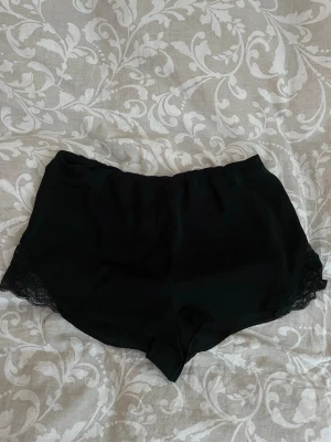 Svarta siden shorts med spetskant - Snygga svarta Pjymas shorts med bred resår i midjan och dekorativ spetskant längs benen. 