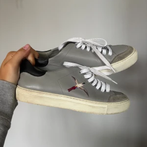 Axel Arigato skor storlek 38  - Snygga grå sneakers från Axel Arigato med vit sula, vita skosnören och svart detalj vid hälen. Rosa lila fågel på sidan och grå mocka framtill. Super fint skick🩷