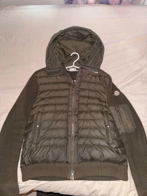 Olivgrön pufferjacka från Moncler - Säljer en olivgrön pufferjacka från Moncler med stickade ärmar och huva. Jackan har två fickor framtill med tryckknappar, ribbade muddar och en Moncler-logga på ärmen. Materialet är en mix av dunfyllning och stickad bomull vilket gör den både varm och snygg.