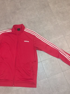 Röd Adidas track jacket - Säljer en klassisk röd Adidas track jacket med vita ränder längs ärmarna och vit Adidas-logga på bröstet. Jackan har dragkedja framtill, hög krage och två fickor. Tillverkad i ett mjukt och stretchigt material som passar perfekt till träning eller streetwear.