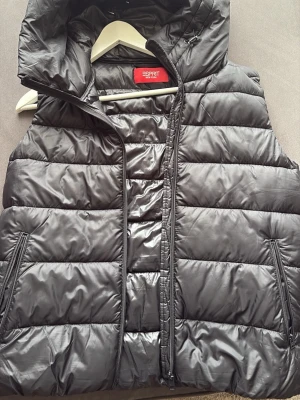 Svart dunväst från Esprit med huva - Snygg svart dunväst från Esprit med quiltad design och dragkedja framtill. Västen har två fickor med dragkedja och en stor, mysig huva. Perfekt för lager-på-lager och håller dig varm under kyliga dagar. Klassisk och stilren look som passar till det mesta.