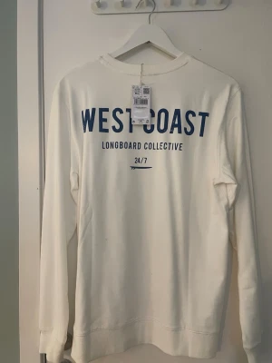 Vit sweatshirt WEST COAST mango - Vit sweatshirt från Mango med trycket 'WEST COAST LONGBOARD COLLECTIVE 24/7' i blått. Tröjan har rund halsringning och långa ärmar. Tillverkad i 100% bomull, perfekt för en avslappnad och cool stil.
