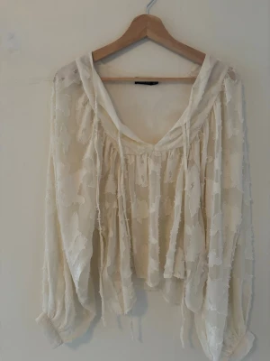 Vit transparent blus med knytband - Säljer en luftig och transparent vit blus med långa ärmar och v-ringning. Blusen har ett subtilt blommönster i tyget och dekorativa knytband vid halsen. Perfekt för lager på lager och ger en romantisk vibe till din outfit.