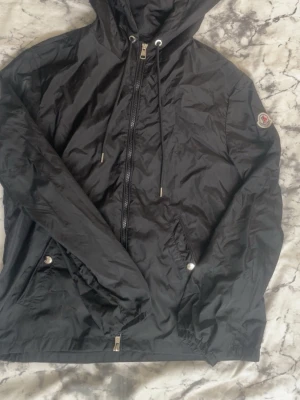 Moncler windbreaker - Snygg svart vindjacka från Moncler med huva och dragsko. Jackan har dragkedja framtill, två fickor med tryckknappar och Moncler-logga på ärmen. Tillverkad i lätt, glansigt material som står emot vind. Perfekt för dig som vill ha en stilren och funktionell jacka.