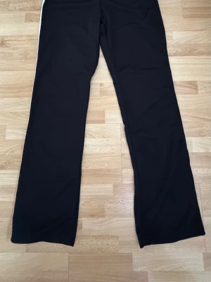  Nya. Svarta mjukisbyxor med vita ränder - Säljer ett par svarta mjukisbyxor med två vita ränder längs sidorna. Byxorna har raka ben och är tillverkade i ett mjukt och stretchigt material, perfekta för en avslappnad stil. Passar dig som gillar sportig och bekväm look. Midjan 85cm/se sista bild lite tråd har lossnat.