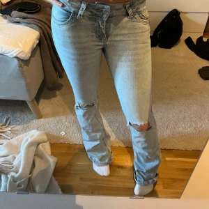 Ljusblå slitna jeans med broderad ficka - Säljer ett par ljusblå jeans med raka ben och slitningar vid knäna. Jeansen har en broderad detalj i rött på bakfickan och klassisk femficksdesign. Materialet är denim och passformen är relaxed. Perfekta för en avslappnad och trendig look.