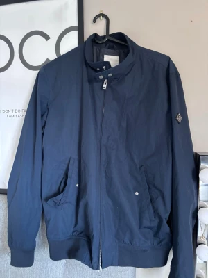 Marinblå bomberjacka J.Lindeberg - Snygg marinblå bomberjacka från J.Lindeberg (som inte säljs längre) med hög krage och dragkedja framtill. Jackan har två snedställda fickor med tryckknappar och ribbade muddar vid ärmslut och nederkant. Märkets logga sitter på vänster ärm. Inga tecken alls på användning, använd någon gång. Skriv gärna för fler bilder! Nypris 3000-4000