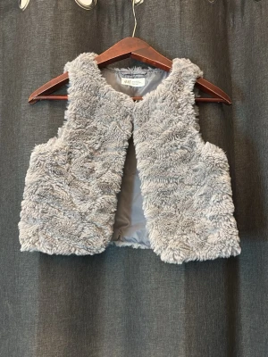 Ljusgrå fuskpälsväst från H&M - Supersnygg ljusgrå fuskpälsväst från H&M med mjukt fluffigt material och helfodrad insida. Västen har en öppen front och kort, boxig passform som ger en trendig look. Perfekt att slänga över en hoodie eller långärmad tröja för extra stilpoäng.